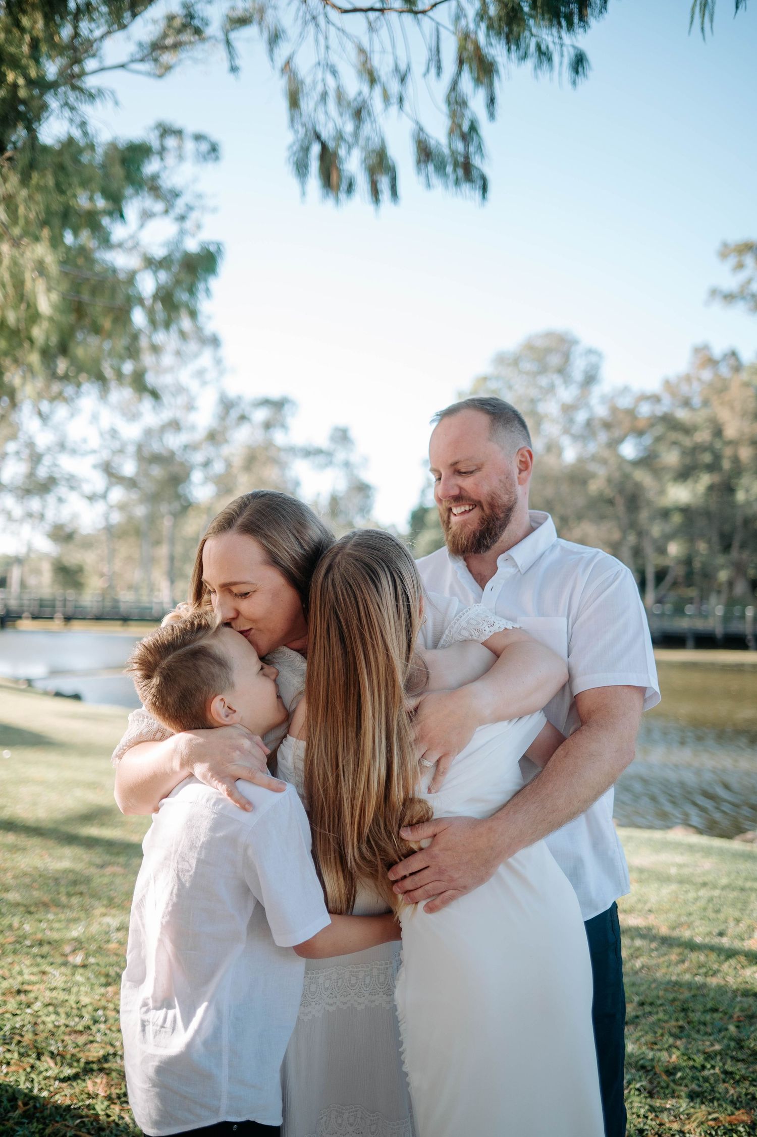 A happy family of four sharing hugs outdoors in a park, dressed in white, with trees and water in the background.