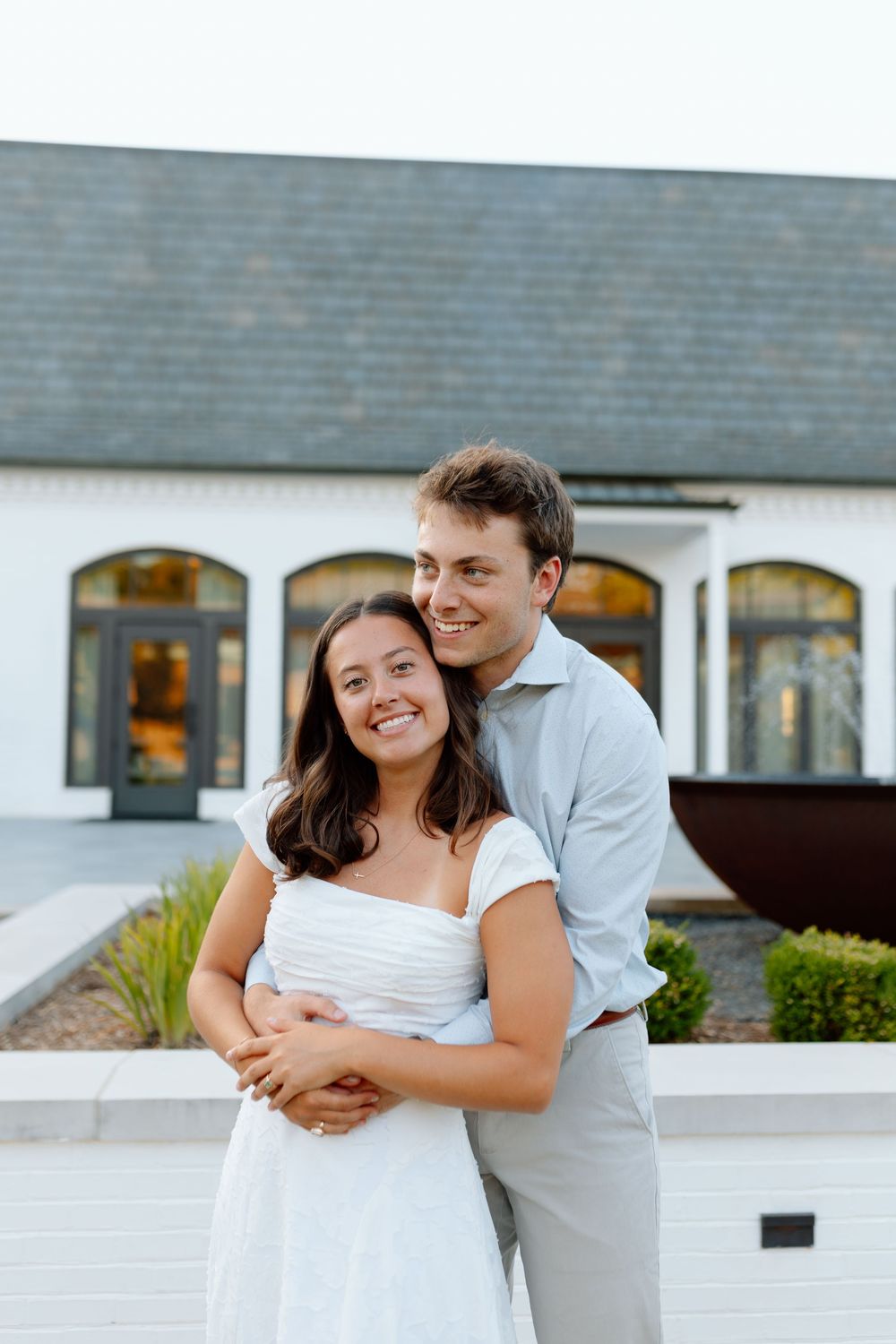 A smiling couple embraces outdoors in front of a white building, woman in white dress, man in light blue shirt.