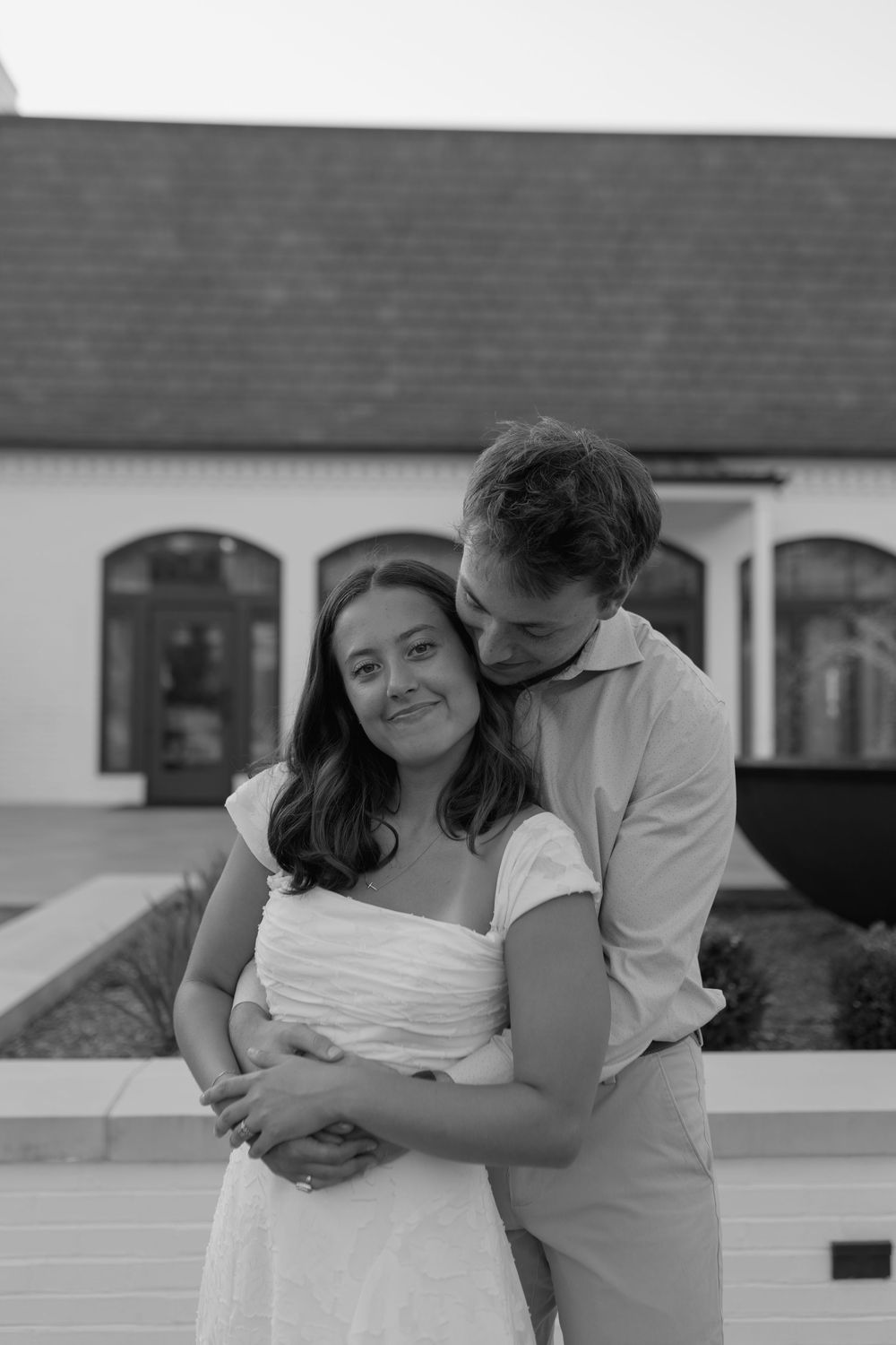 Black and white photo of a couple embracing outdoors, man kissing womans head, white dress, building background.
