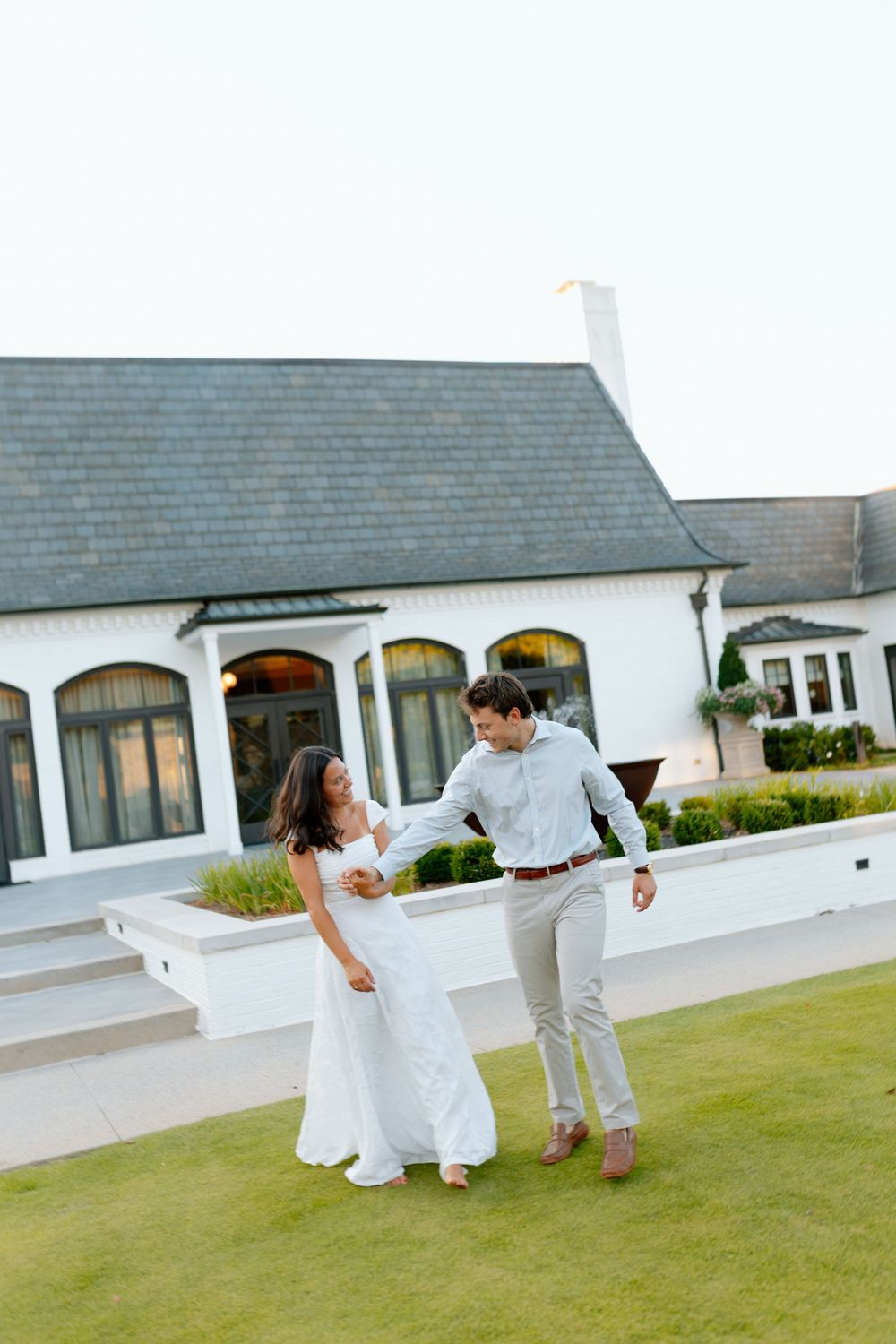 Couple in white dress and light blue shirt dancing on green lawn in front of elegant white building.
