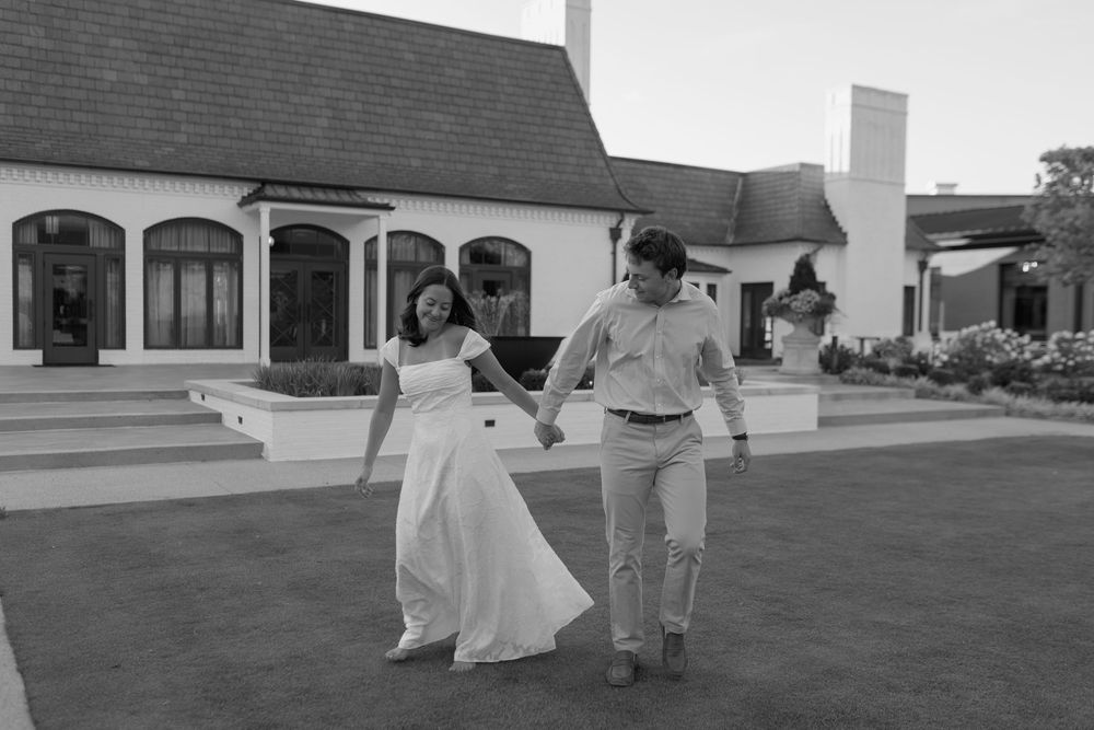 Black and white photo of a bride and groom holding hands walking on a lawn outside an elegant white wedding venue.