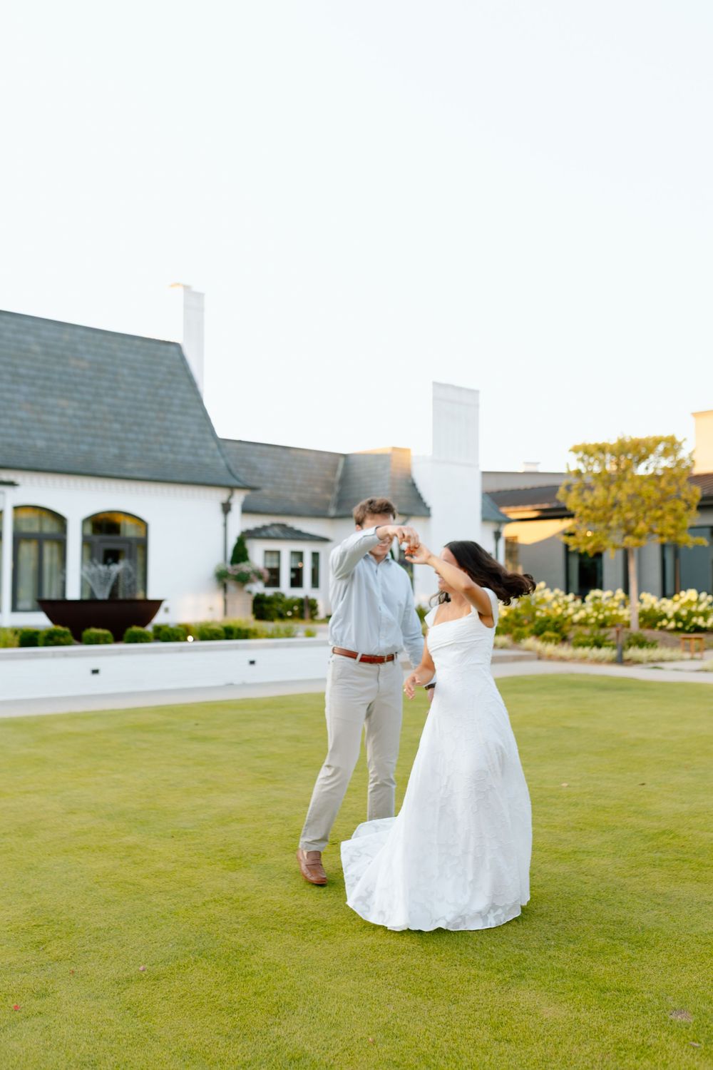 A bride and groom dance together on a green lawn outside a white estate during their outdoor wedding celebration.