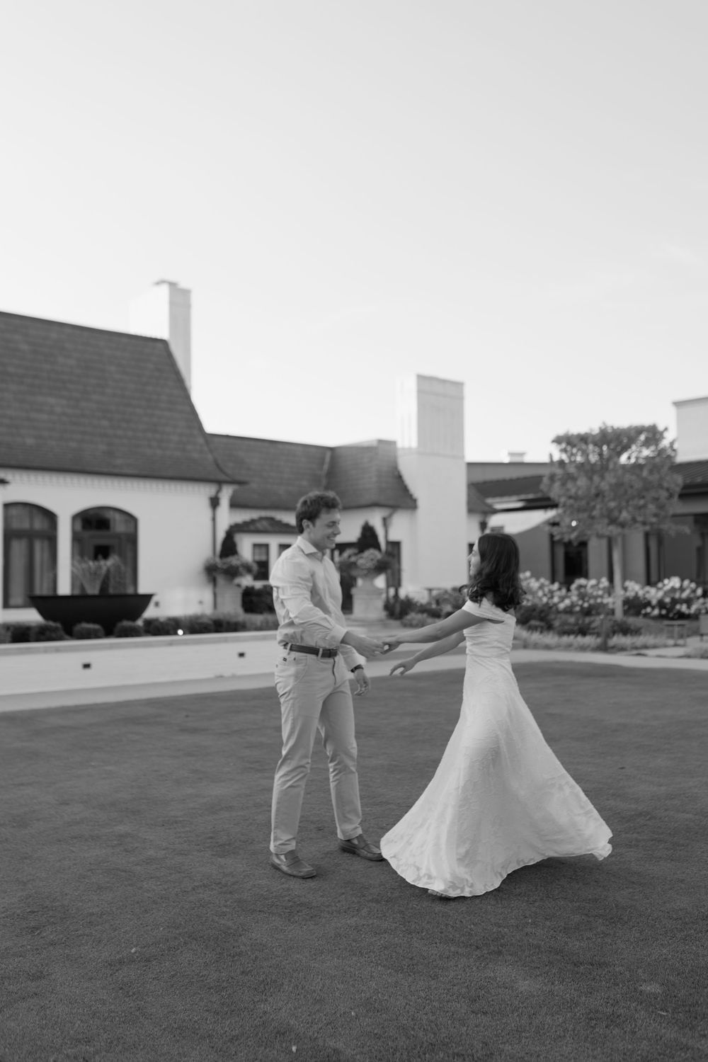 Black and white photo of a bride and groom dancing outdoors on a lawn with elegant white buildings in the background.