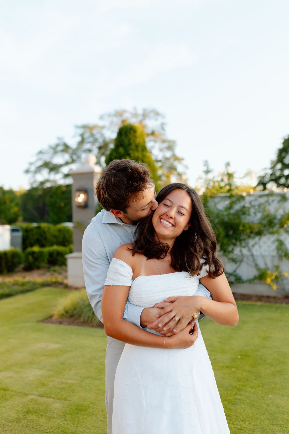 Couple embracing outdoors, man kissing woman's cheek, she smiles in white off-shoulder dress on green lawn.