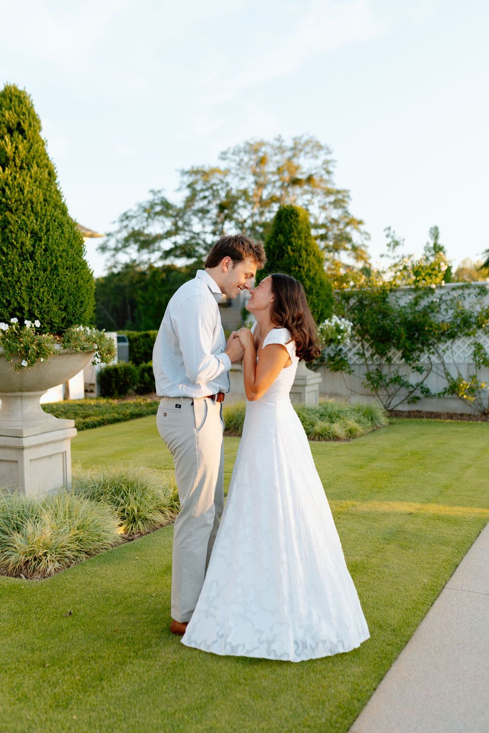 Bride and groom share a romantic kiss in a lush garden, bride in white gown, groom in light gray suit.