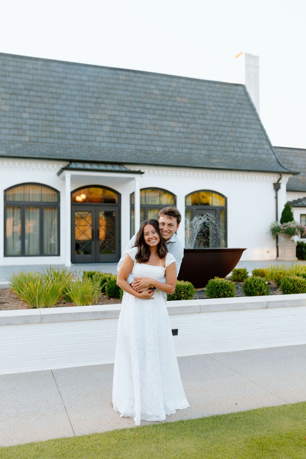 A couple embraces in front of an elegant white venue, the woman wearing a flowing white dress, smiling together outdoors.