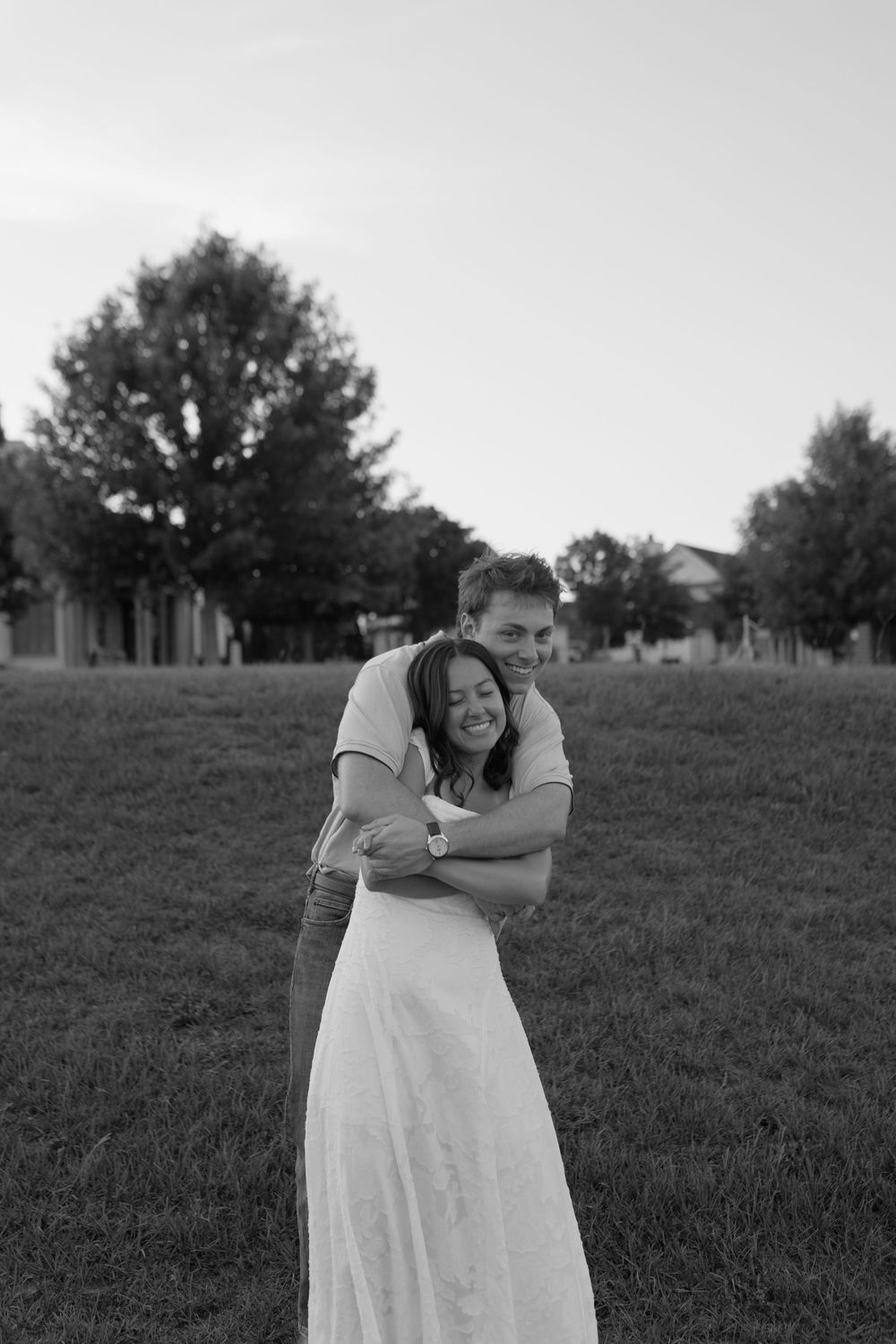 Black and white photo of a couple embracing and smiling in a grassy field with trees in the background.