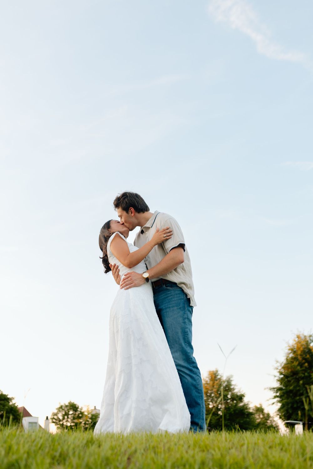 Couple sharing a romantic kiss outdoors on green grass, woman in white dress, man in jeans, blue sky background.