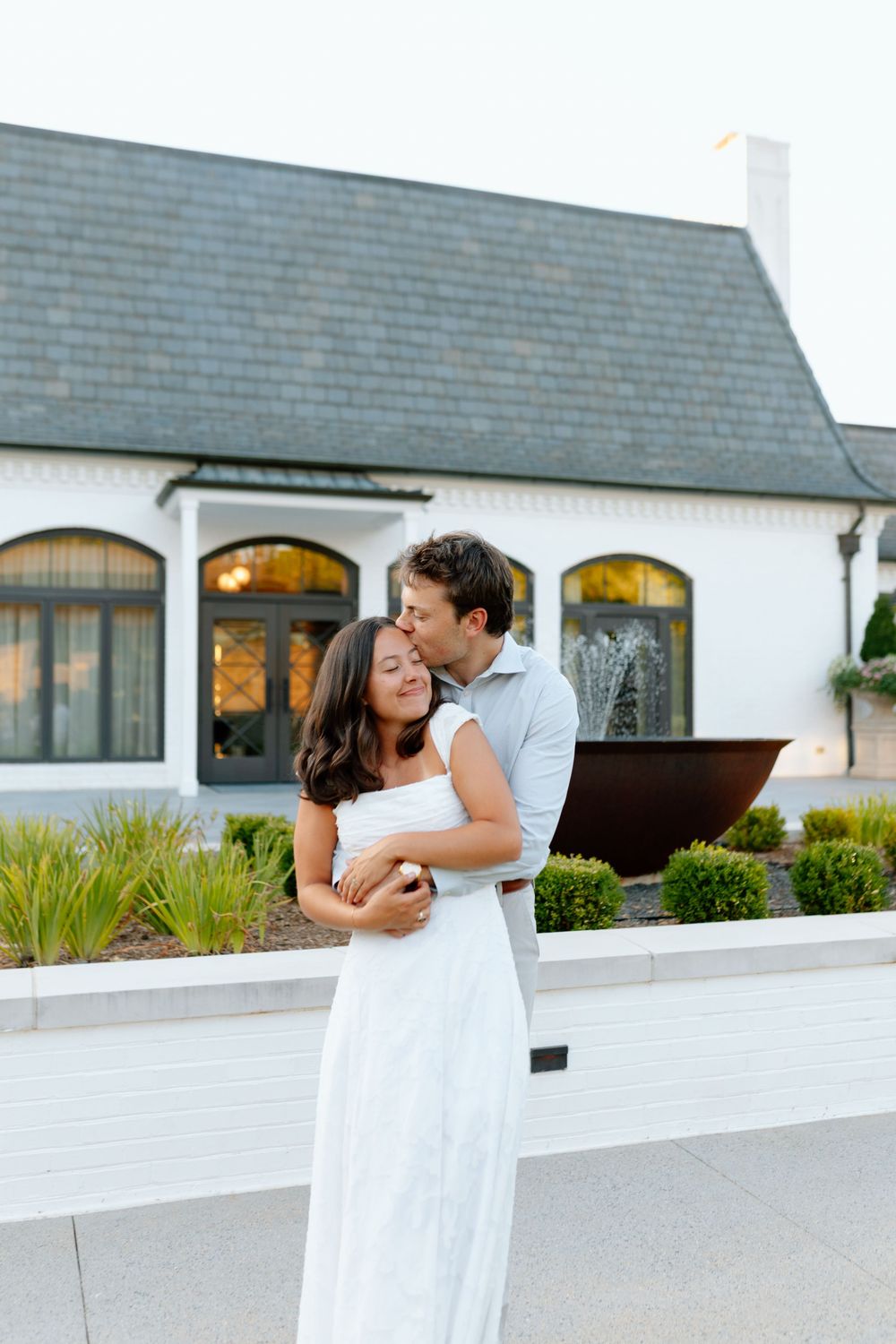 A couple embraces in front of a white house, the man kissing the woman's cheek as she smiles in a white dress.