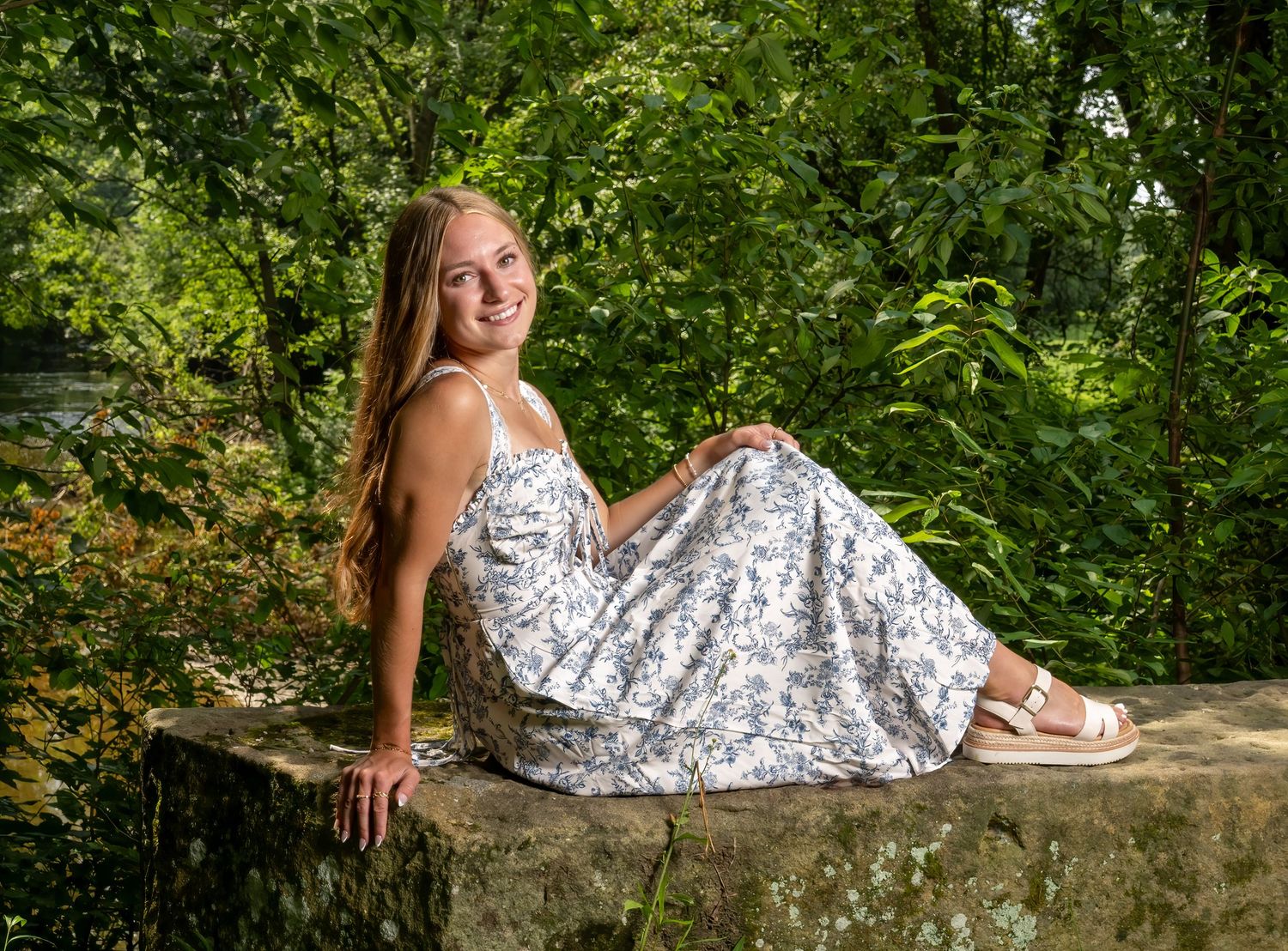 Young woman in floral sundress sitting on stone wall smiling outdoors surrounded by lush green trees.