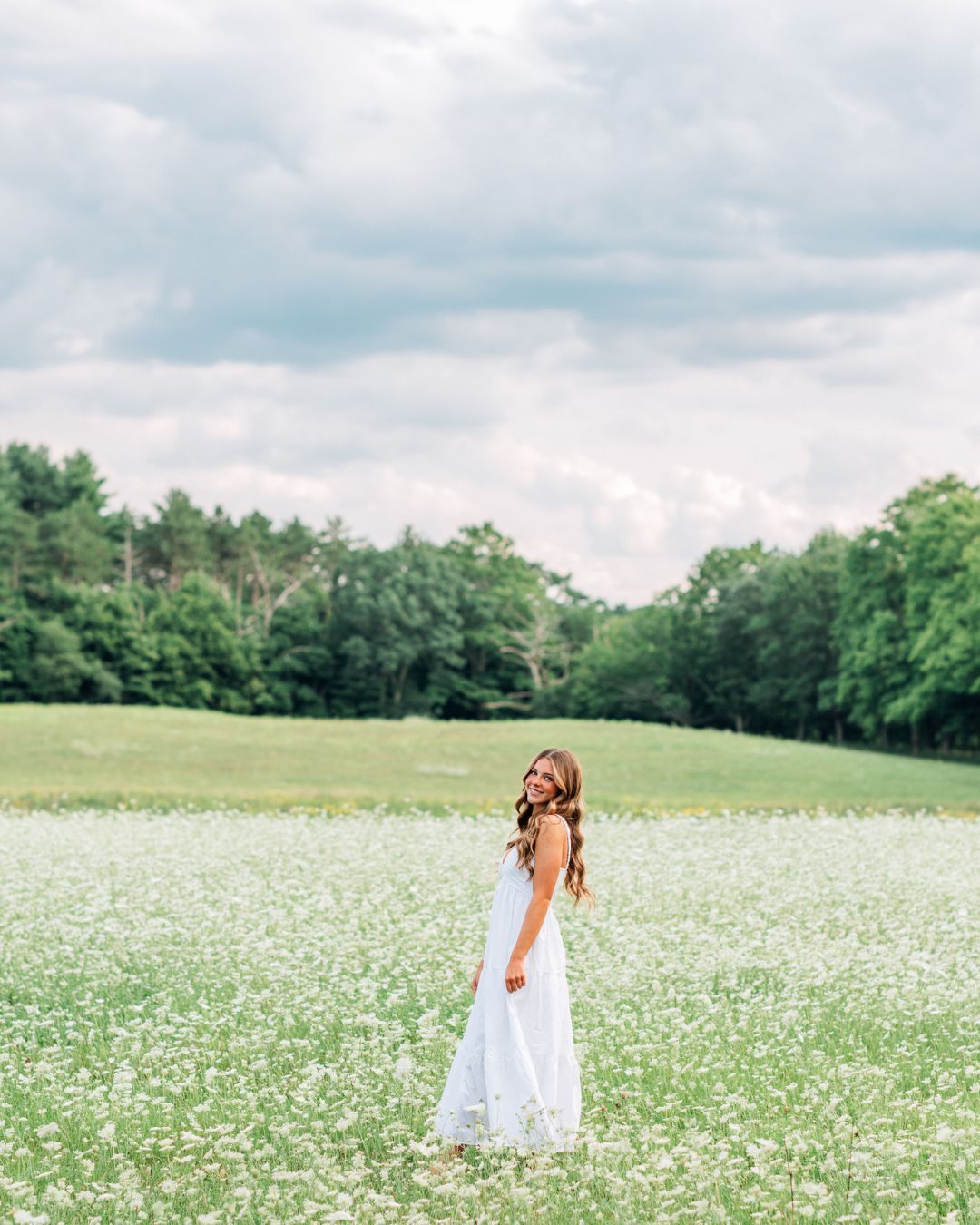 A young woman in a white maxi dress stands in a blooming wildflower field with lush green trees under a cloudy sky.