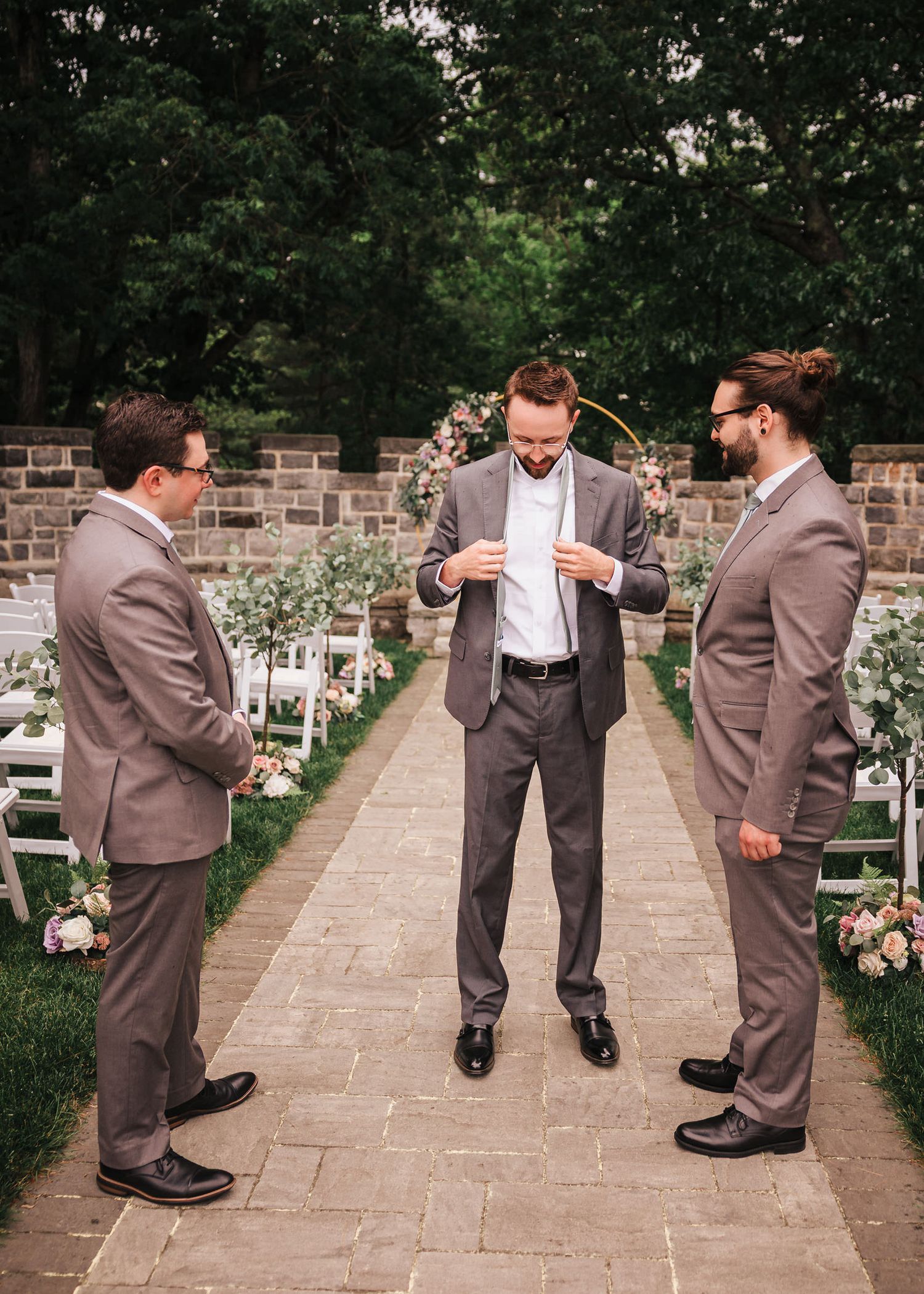 Groom adjusting his tie in the ceremony aisle at Searles Castle, flanked by two groomsmen in matching gray suits.