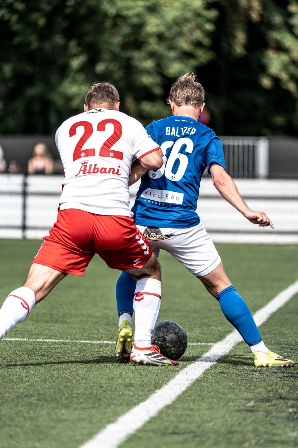 Two soccer players compete for the ball, player 22 in white and red vs player in blue Balzer jersey on green turf.