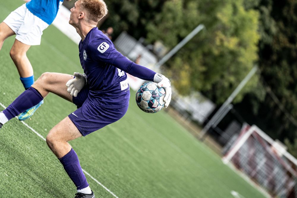 A goalkeeper in purple uniform kicks a soccer ball during a competitive match on a green turf field.