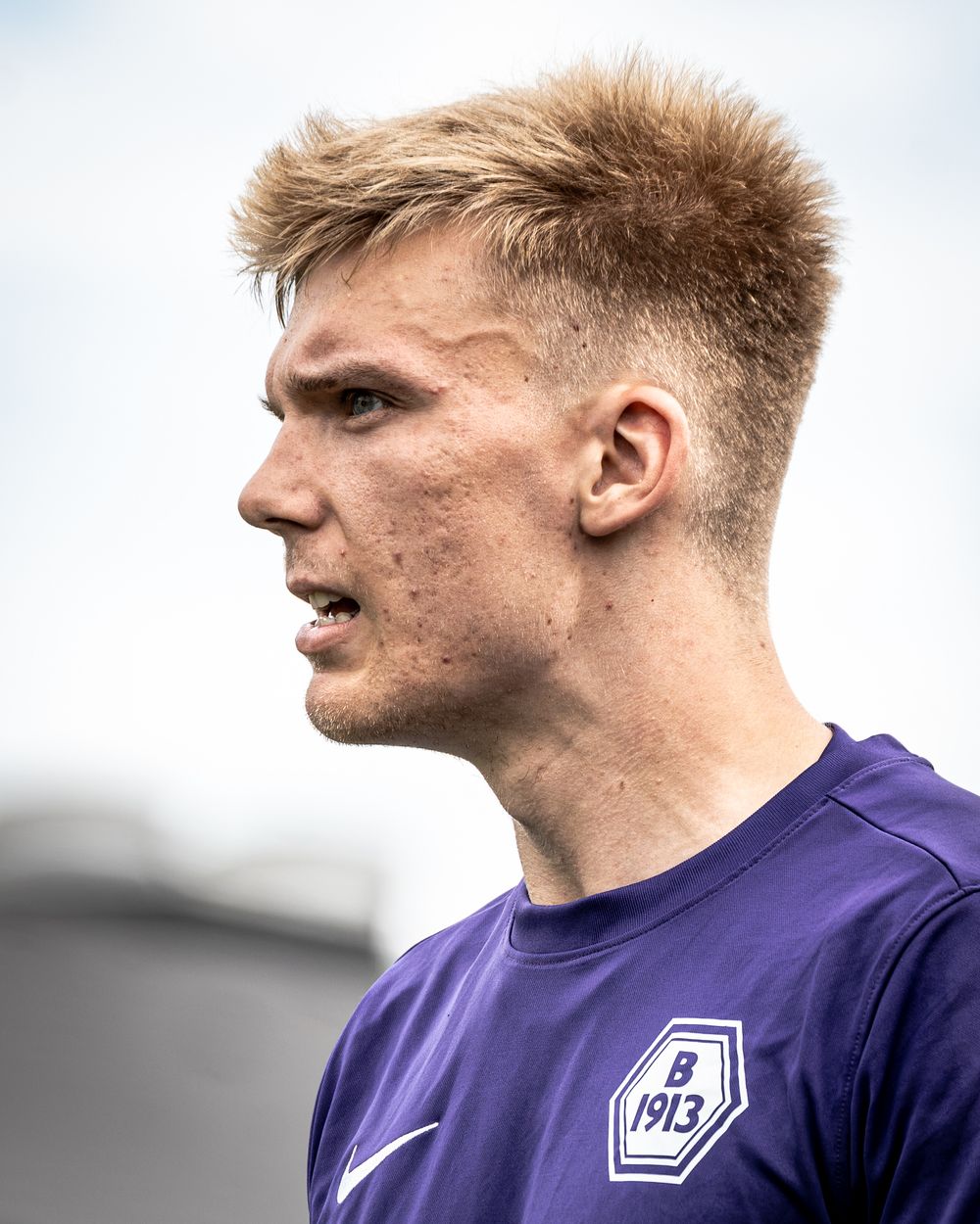 Young male soccer player in purple B1913 jersey with blonde hair, shown in close-up side profile outdoors.