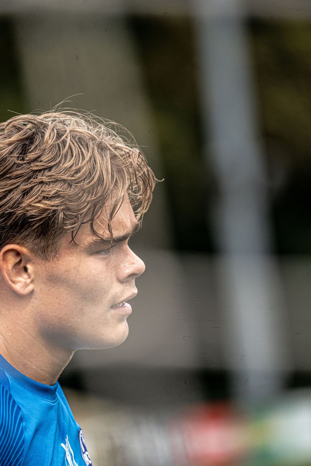 Young male athlete with wet hair in profile view, wearing a blue jersey during an outdoor sports event.