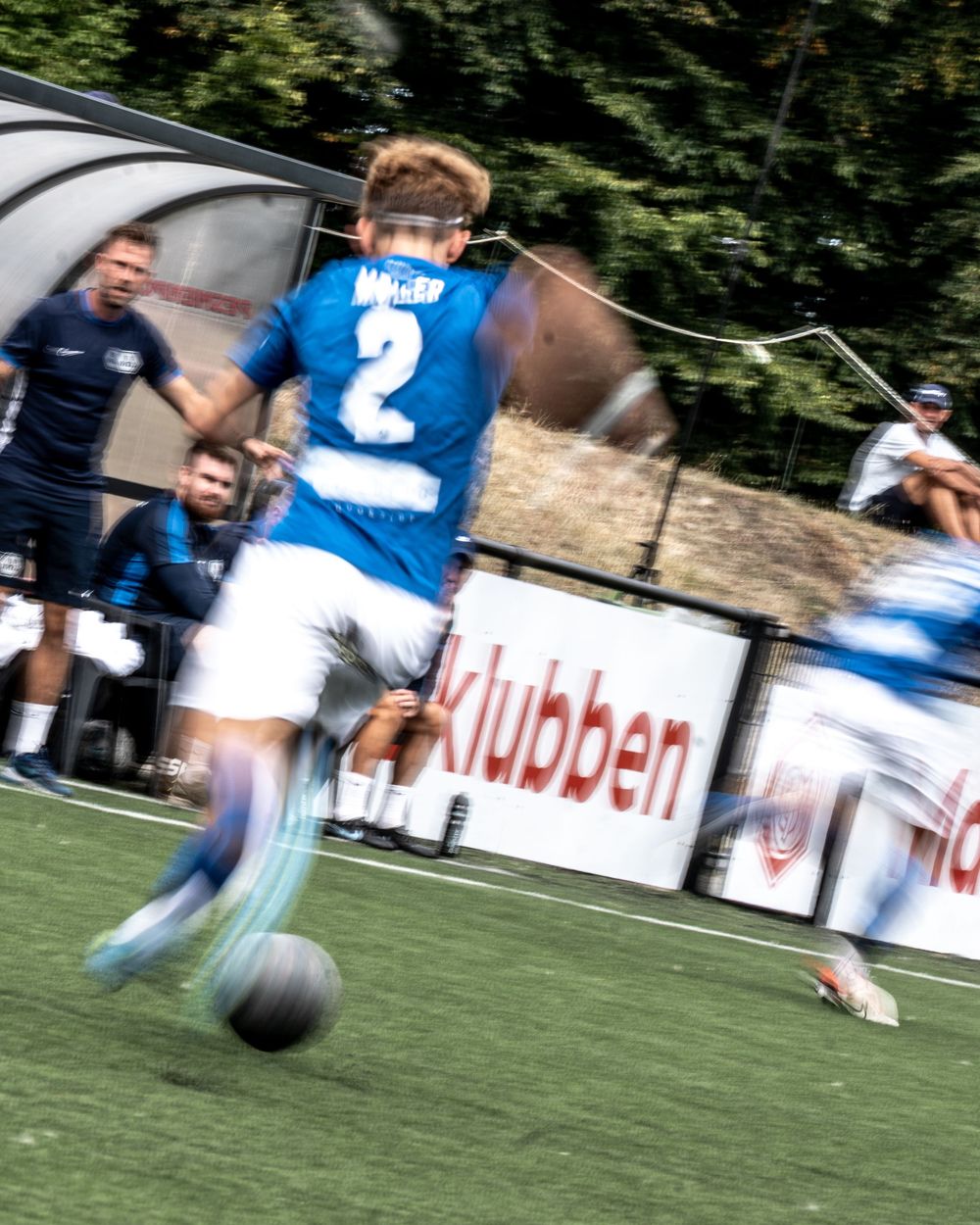 Soccer player wearing blue number 2 jersey dribbles ball during competitive match with motion blur effect.