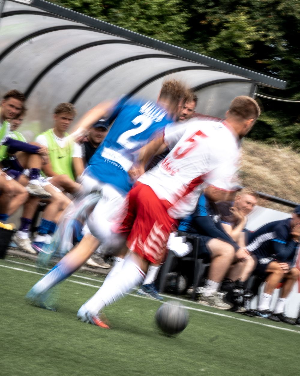 Two soccer players in blue and red uniforms battle for the ball near the sideline bench during a competitive match.