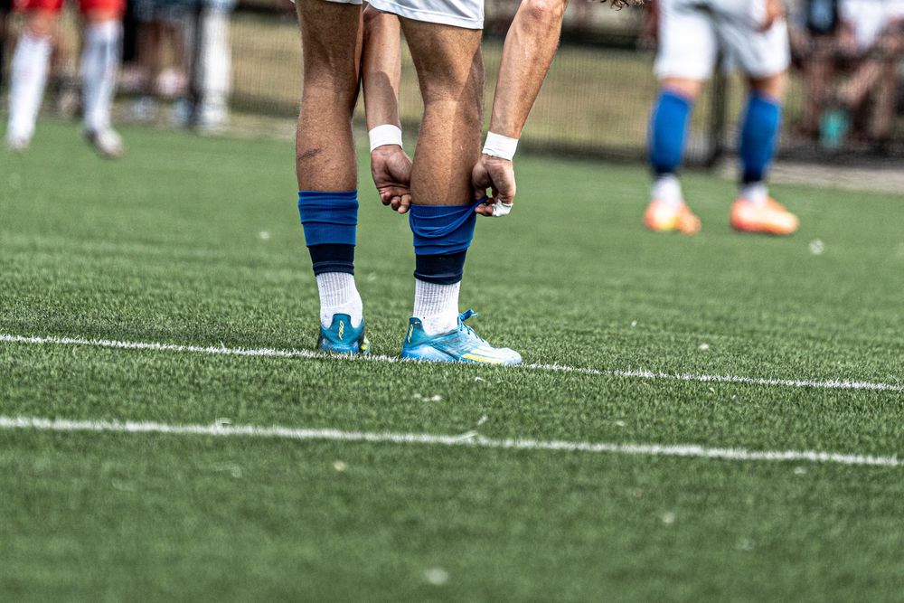 Soccer player adjusting blue cleats on green turf field during a match.