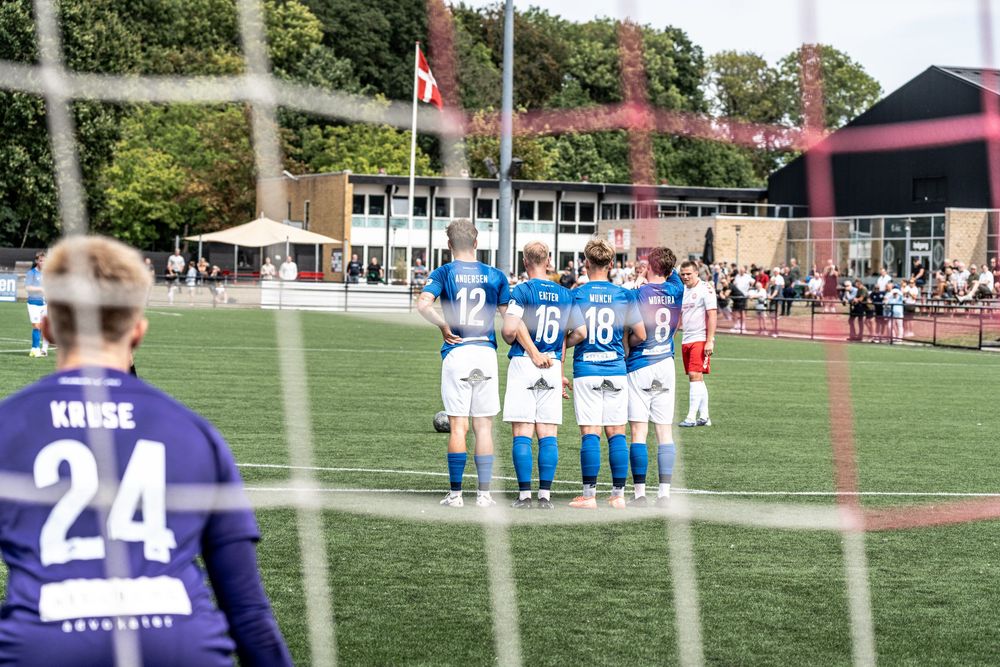 Female soccer players in blue jerseys huddle on a green field during a match, with a goalkeeper wearing number 24 nearby.