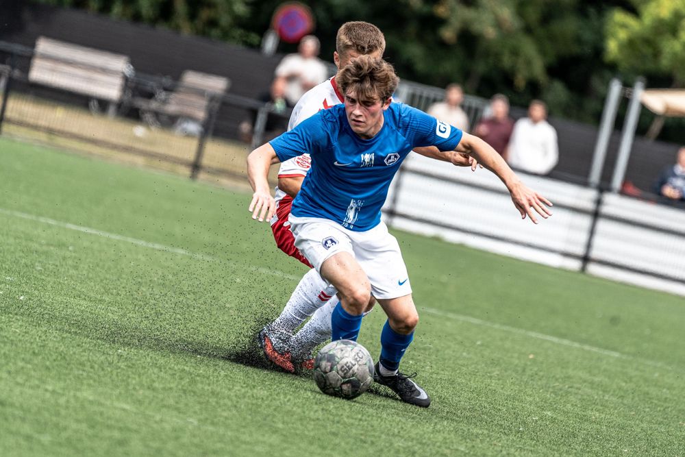 Two youth soccer players compete for the ball during a match on a green turf field, one in blue and one in red.