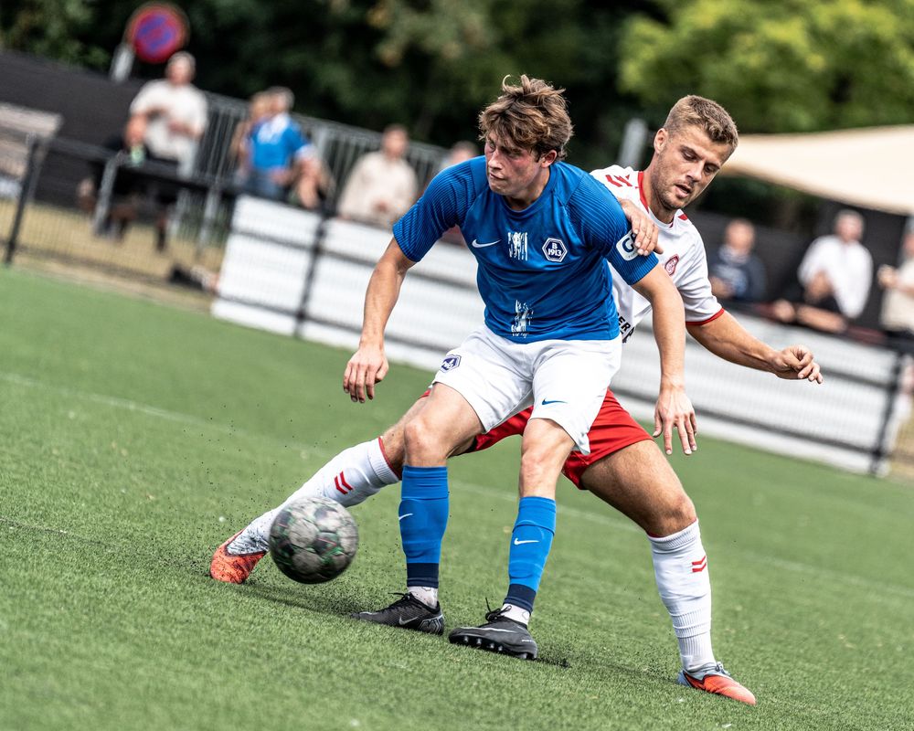 Two soccer players compete for the ball during an outdoor match, one in blue and one in white and red uniforms.