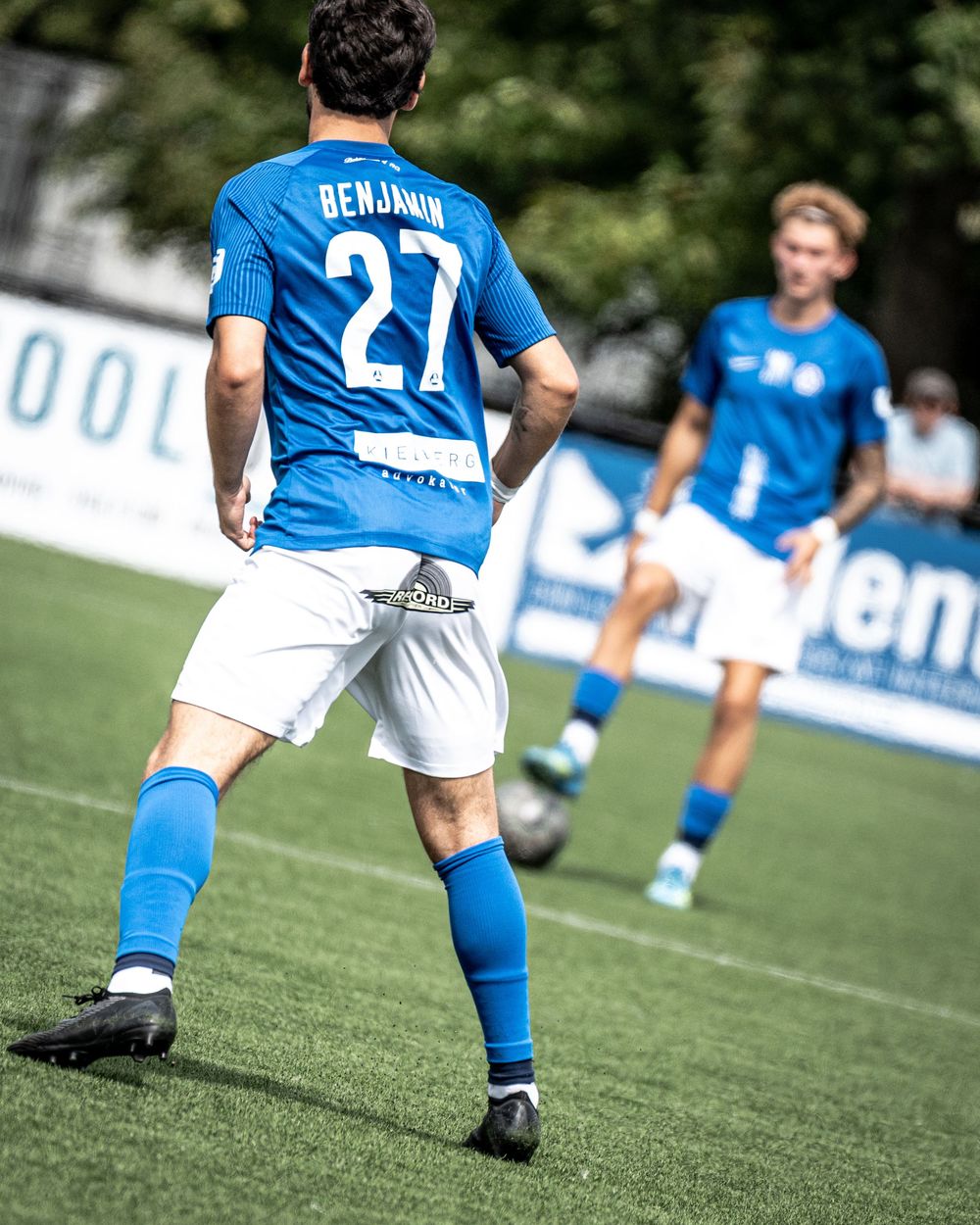 Soccer player Benjamin wearing jersey number 27 in blue and white uniform dribbles ball on green field during match.