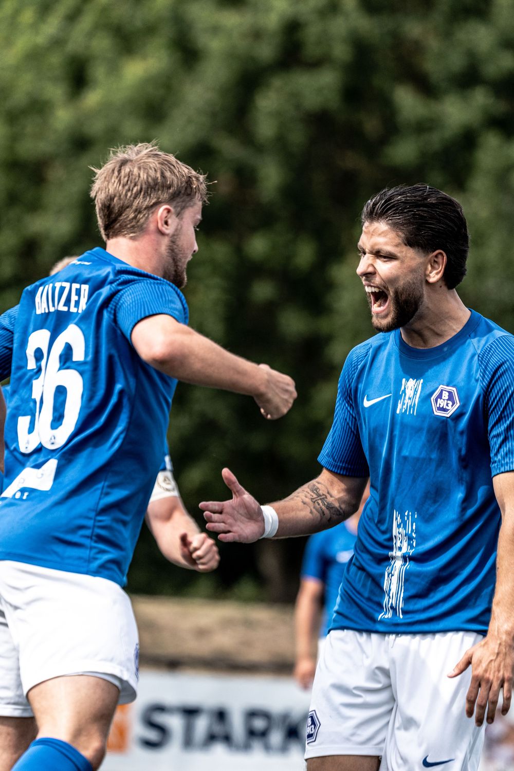 Two soccer players in blue jerseys celebrate a goal on the field, with player number 36 fist-pumping in excitement.