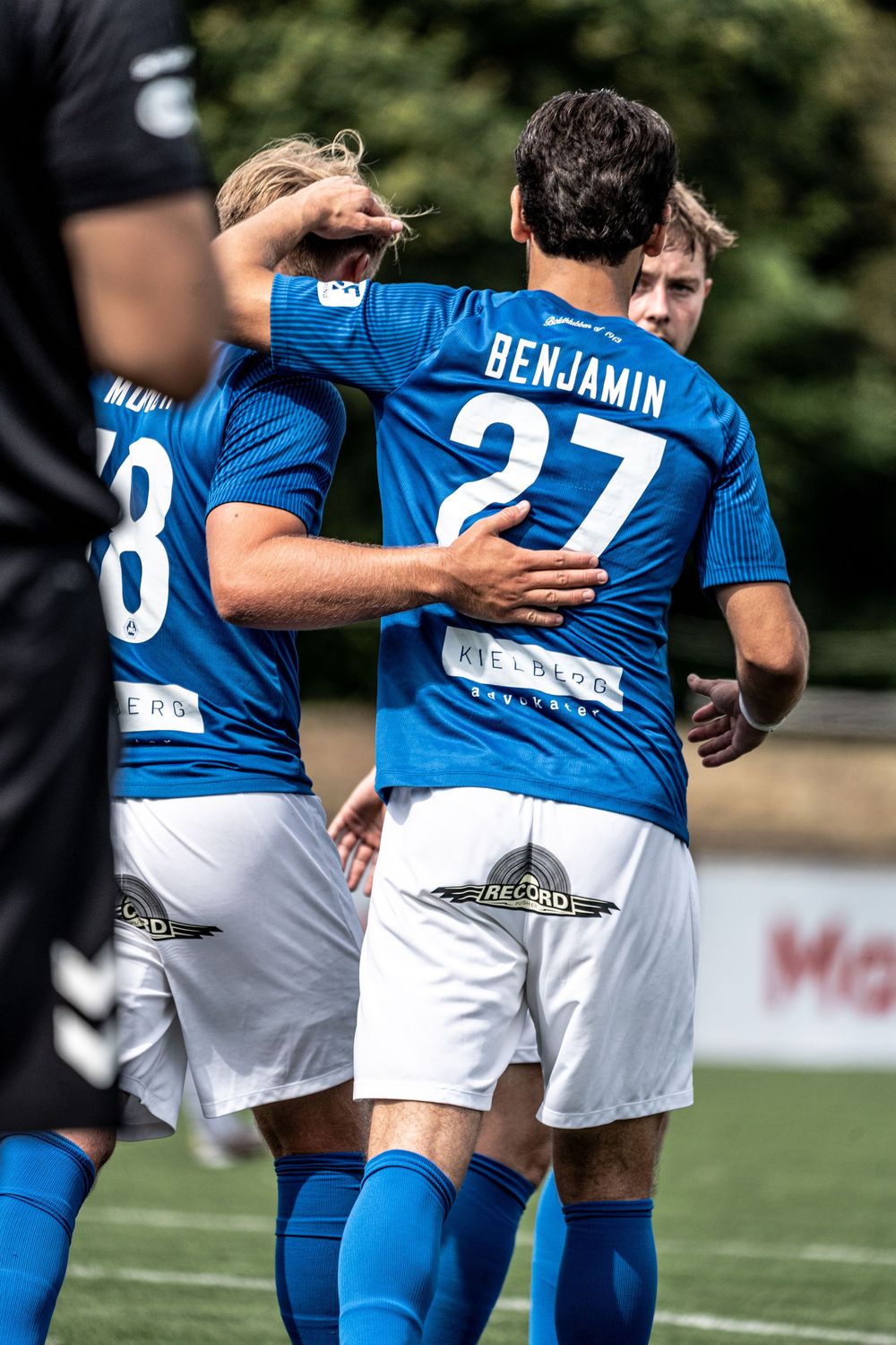 Soccer players in blue jerseys celebrate on field, player number 27 Benjamin embraced by teammate during match.