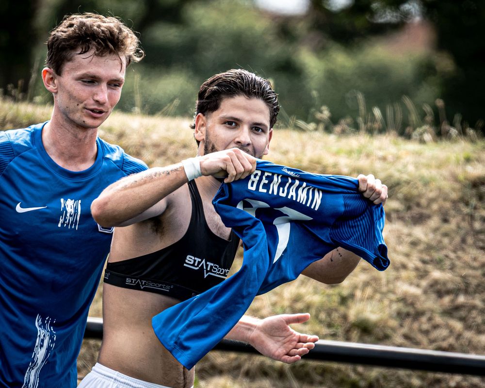 Two soccer players in blue Nike jerseys, one holding up a shirt reading Benjamin after a match outdoors.