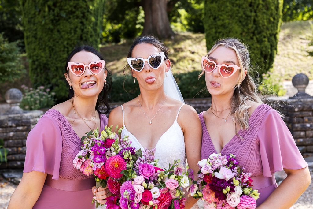 Bride and two bridesmaids in pink dresses wearing heart-shaped sunglasses, holding colorful bouquets outdoors.