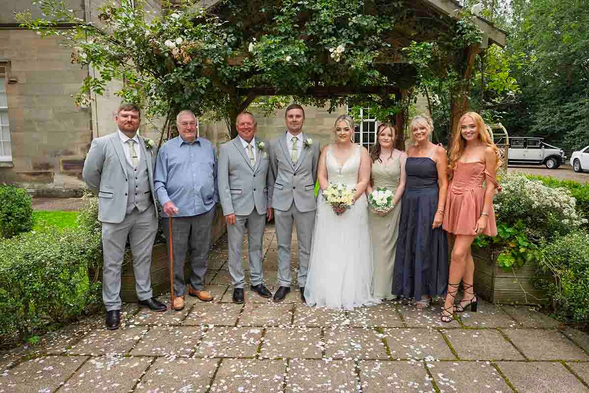 Wedding party posing outdoors at a stone venue, featuring bride and groom with family in formal attire.