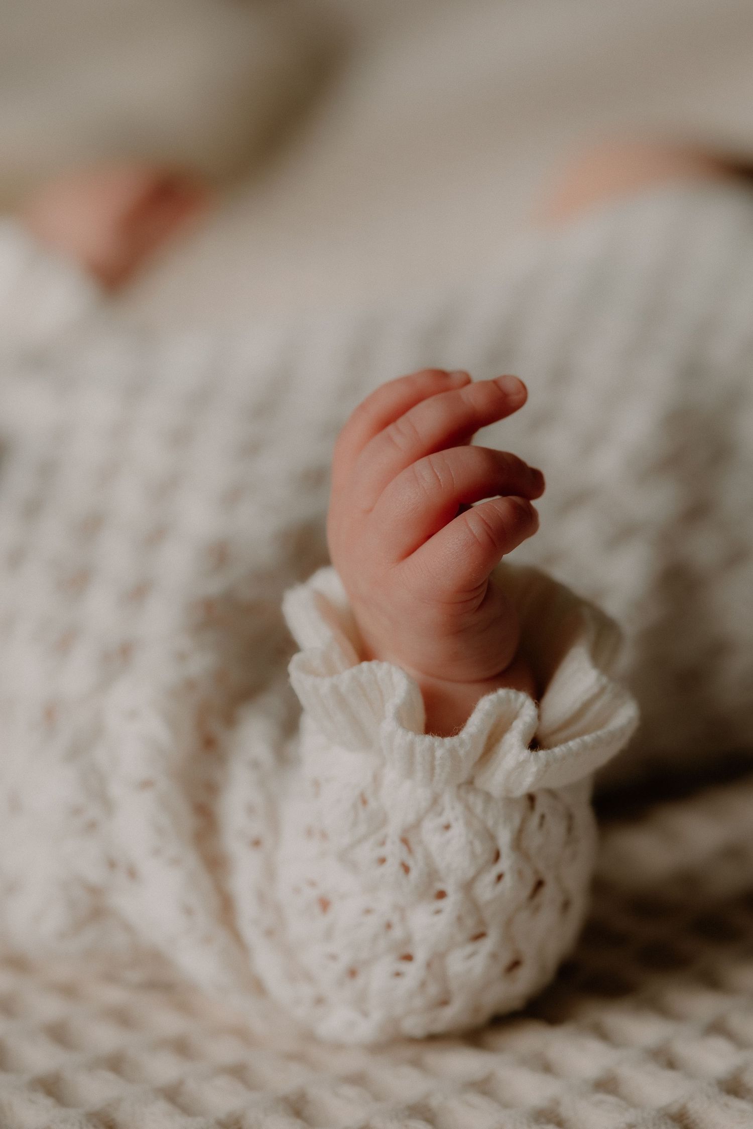 Close-up of a newborn baby's tiny hand peeking out from a white knit sweater sleeve on a soft blanket.