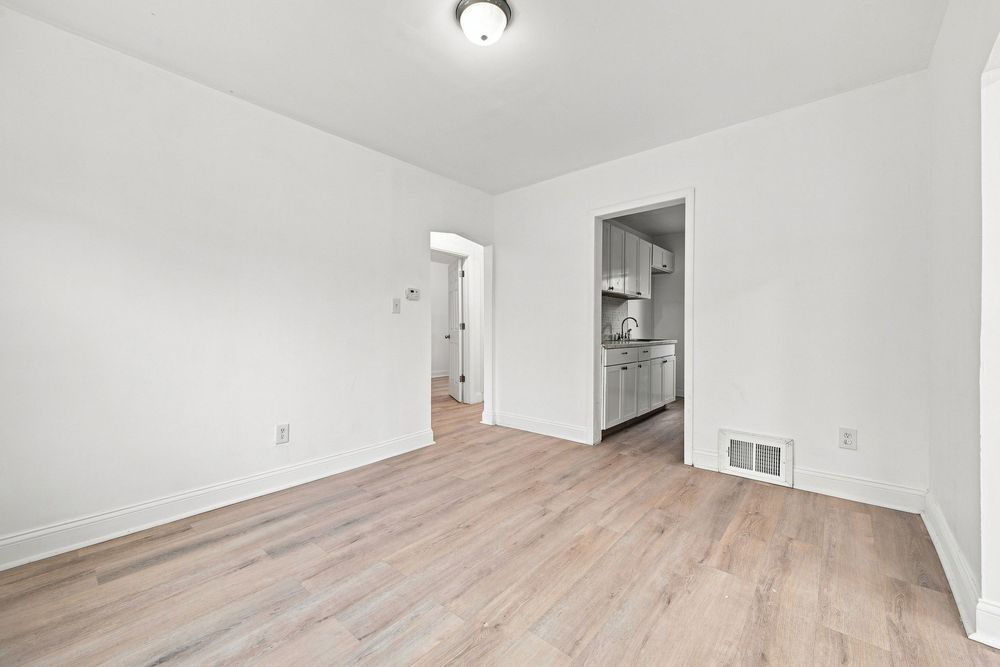 Empty room with light hardwood floors, white walls, and a view into a kitchen through an open doorway.