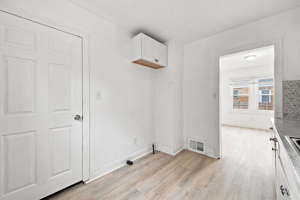 Bright white room with light wood floors, a wall-mounted shelf, floor vent, and doorway leading to a sunlit space.