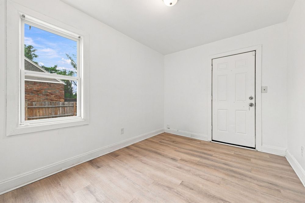 Bright empty room with light wood floors, white walls, a window with backyard view, and a white exterior door.