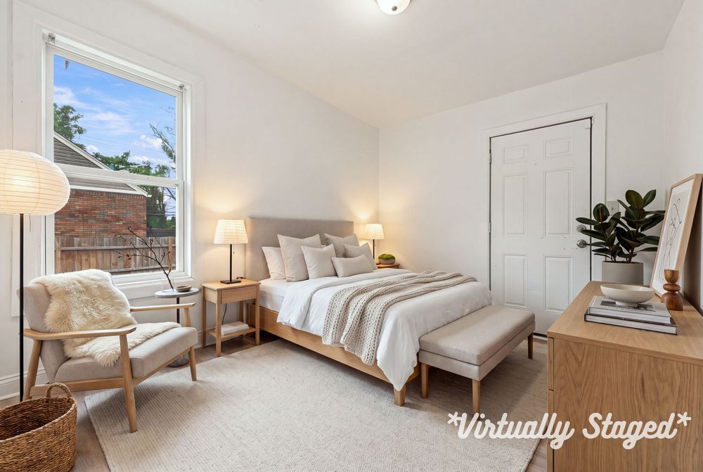 Virtually staged bedroom with neutral tones, wood furniture, cozy armchair, and natural light from a large window.