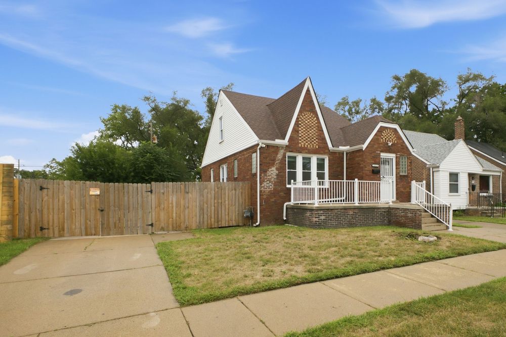 Brick bungalow home with white trim, covered porch, wood fence, and lush green lawn under a clear blue sky.