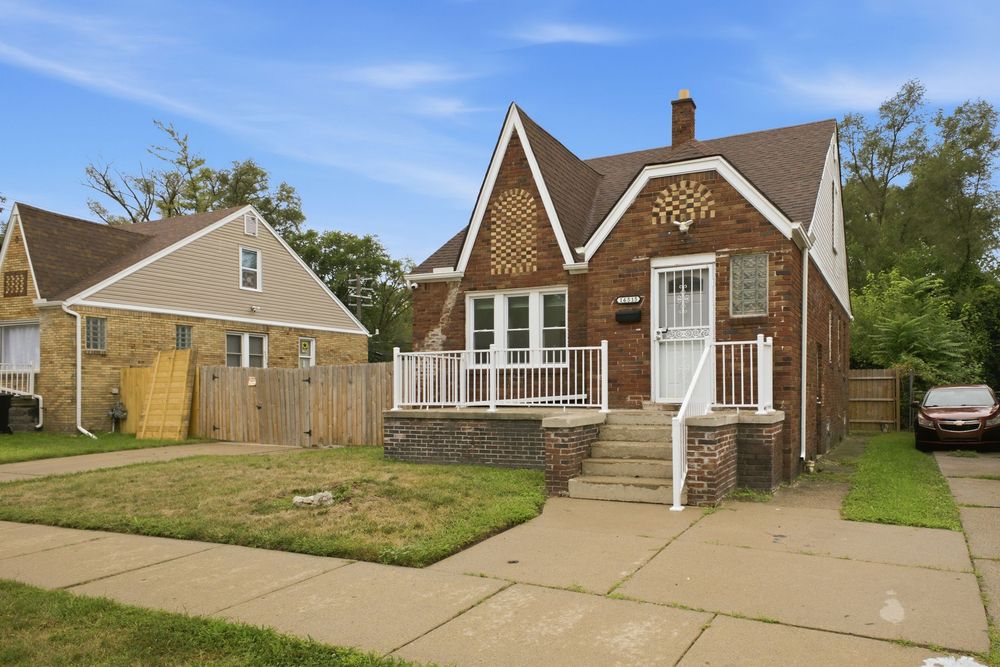 Brick bungalow home with white porch railings, manicured lawn, and wooden fence beside neighboring yellow house.