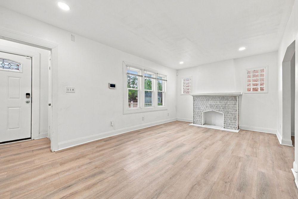 Bright living room with light hardwood floors, white walls, brick fireplace, and natural light from a window.
