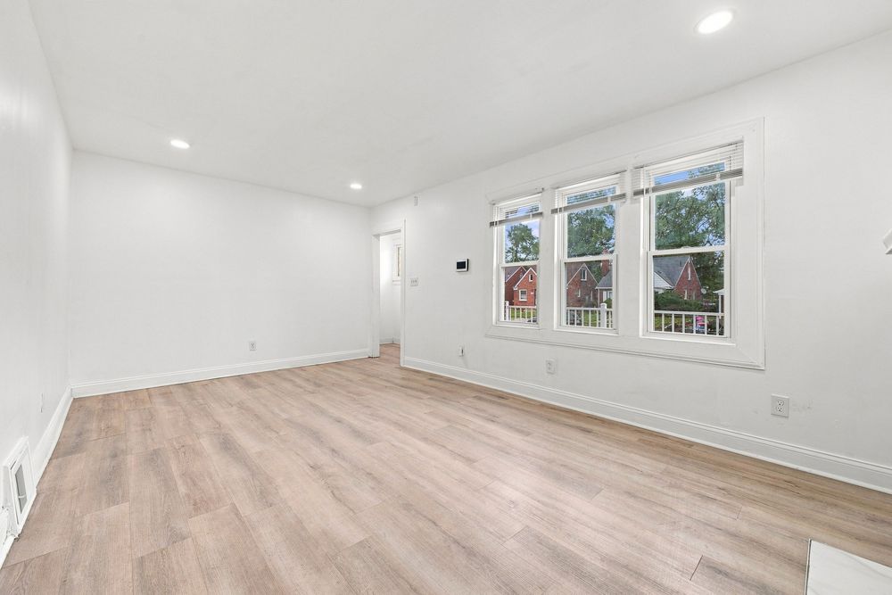 Bright empty living room with light hardwood floors, white walls, recessed lighting, and a large window with natural light.