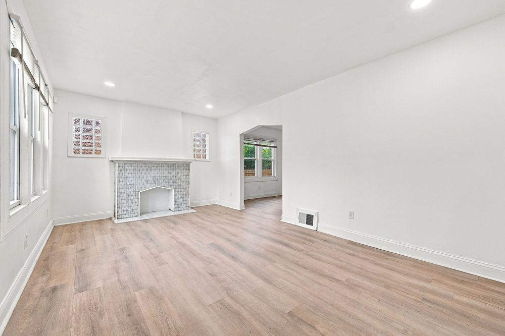 Bright empty living room with hardwood floors, white brick fireplace, recessed lighting, and doorway leading to sunlit room.
