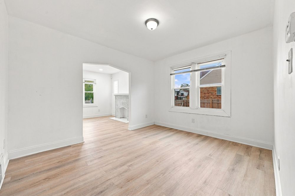 Bright empty room with light hardwood floors, white walls, and a window overlooking a neighborhood.