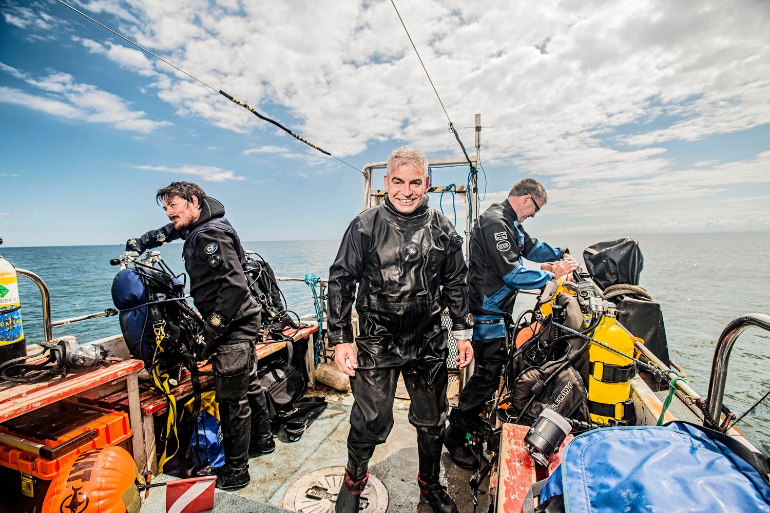 Suffolk Wildlife Ocean Protection Team. Scuba divers in wetsuits prepare to dive on a boat deck, with the ocean and blue sky behind them.
