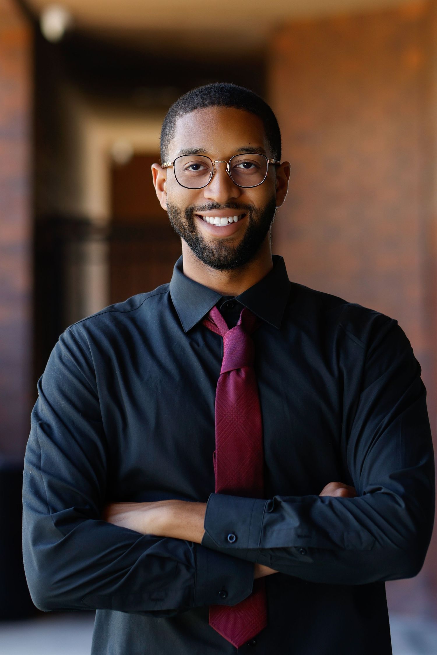 Smiling Black businessman in black shirt and red tie with crossed arms wearing glasses in front of brick wall.