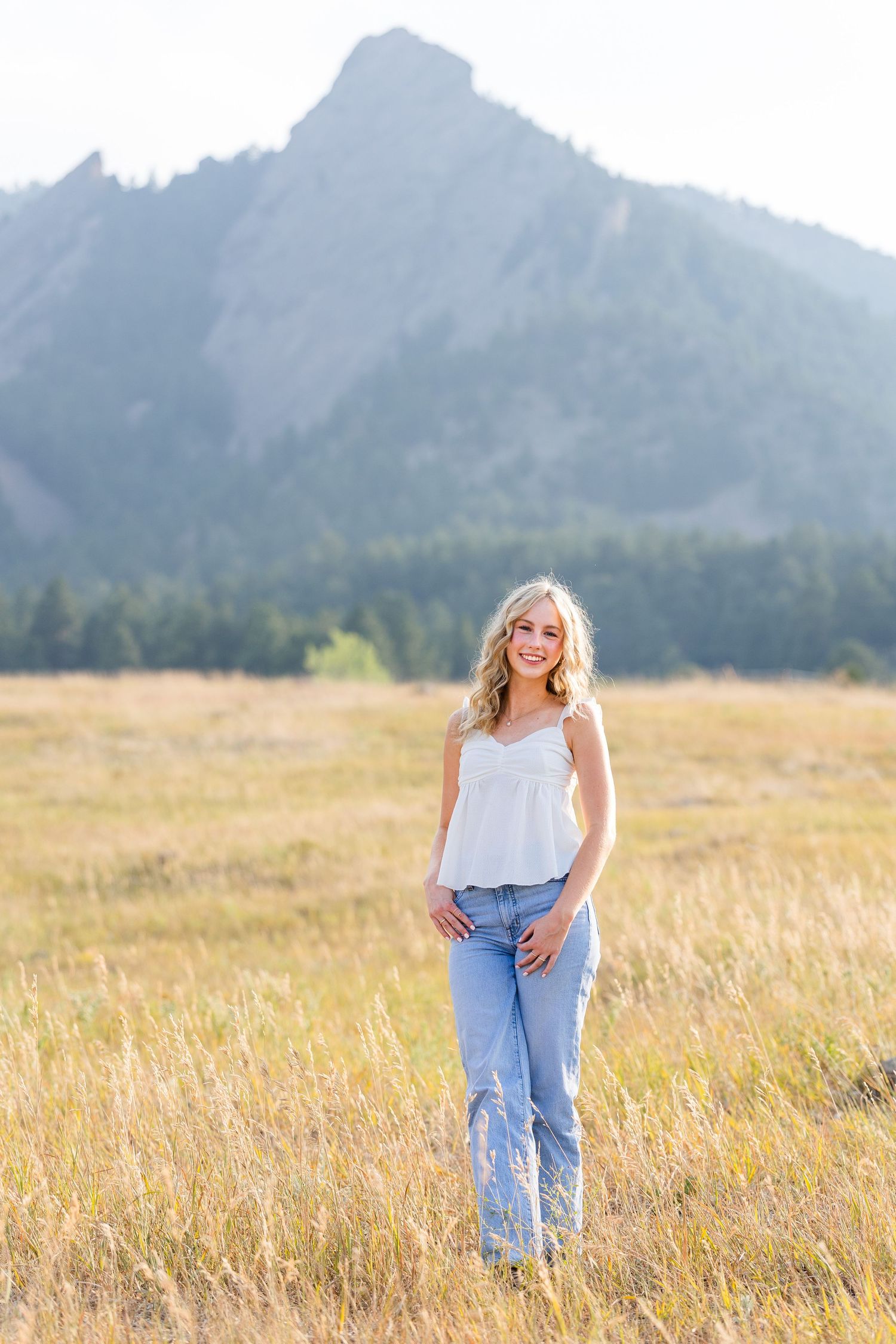 Smiling blonde teen girl in white tank and jeans poses in golden meadow with rocky mountain backdrop.