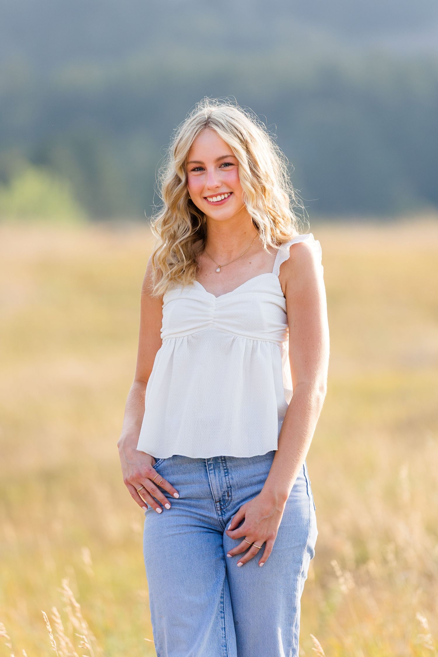 Smiling blonde teen girl in white camisole and jeans poses in a sunny golden meadow with mountains behind her.