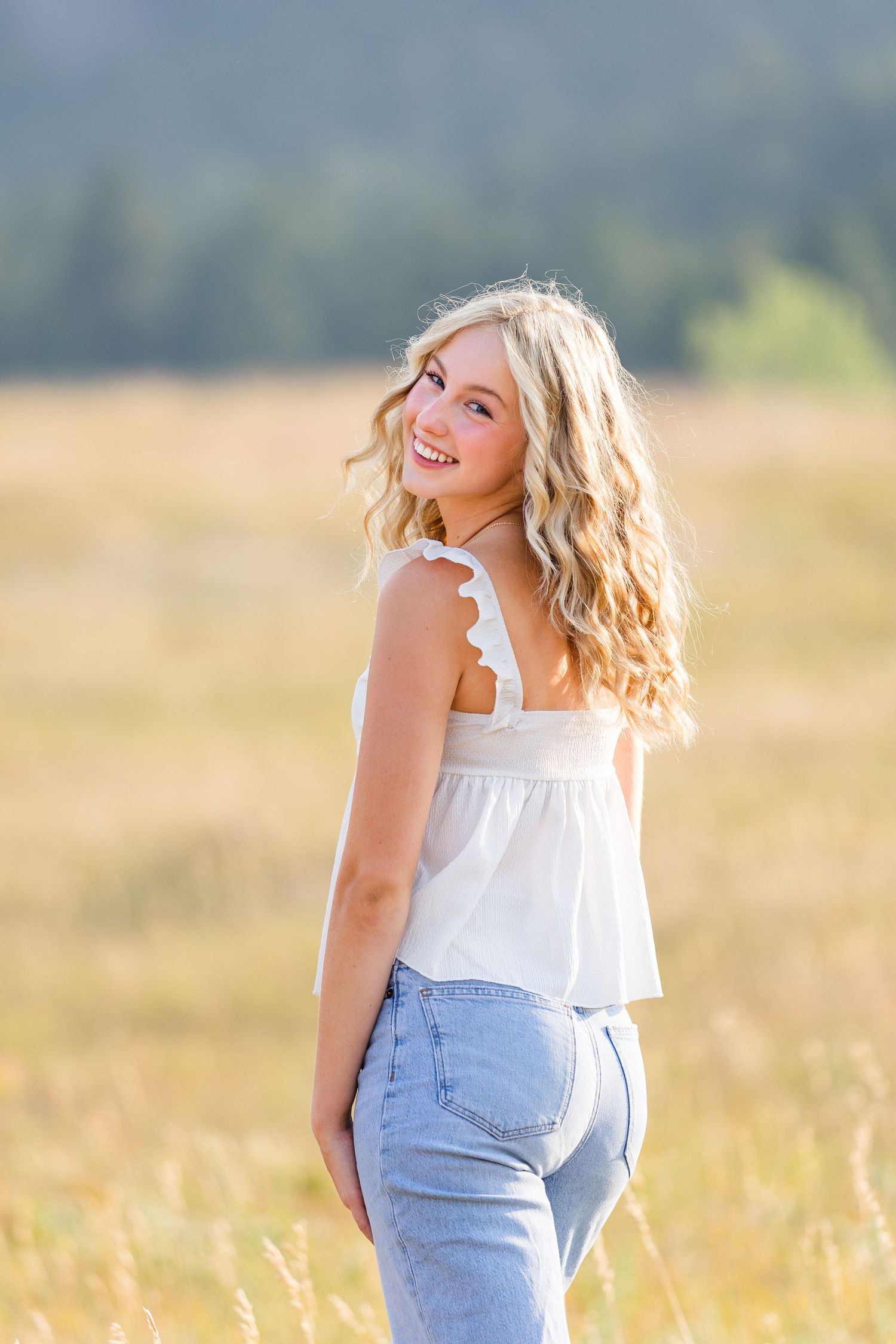 Smiling blonde teen girl in white top and jeans poses in golden field with mountain backdrop.