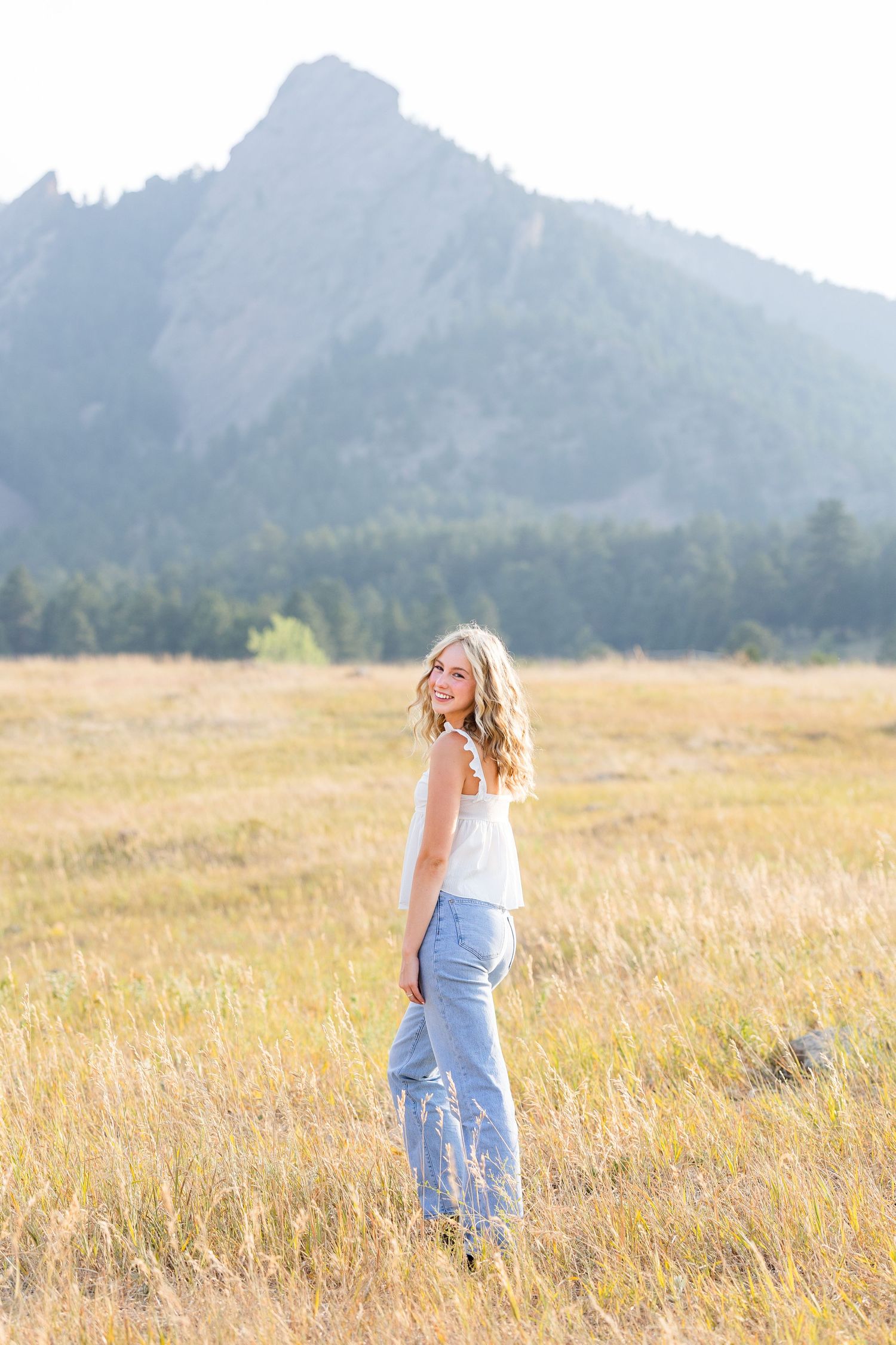 A smiling blonde woman in white top and jeans stands in a golden meadow with the Flatirons mountains in the background.