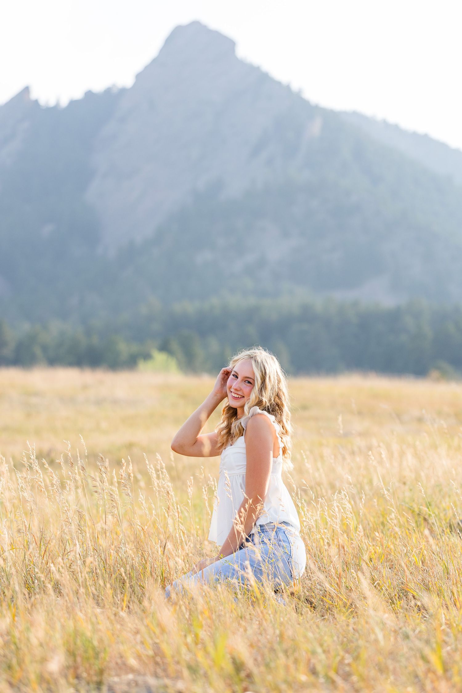 Smiling blonde woman in white top and jeans sitting in golden field with mountain backdrop.